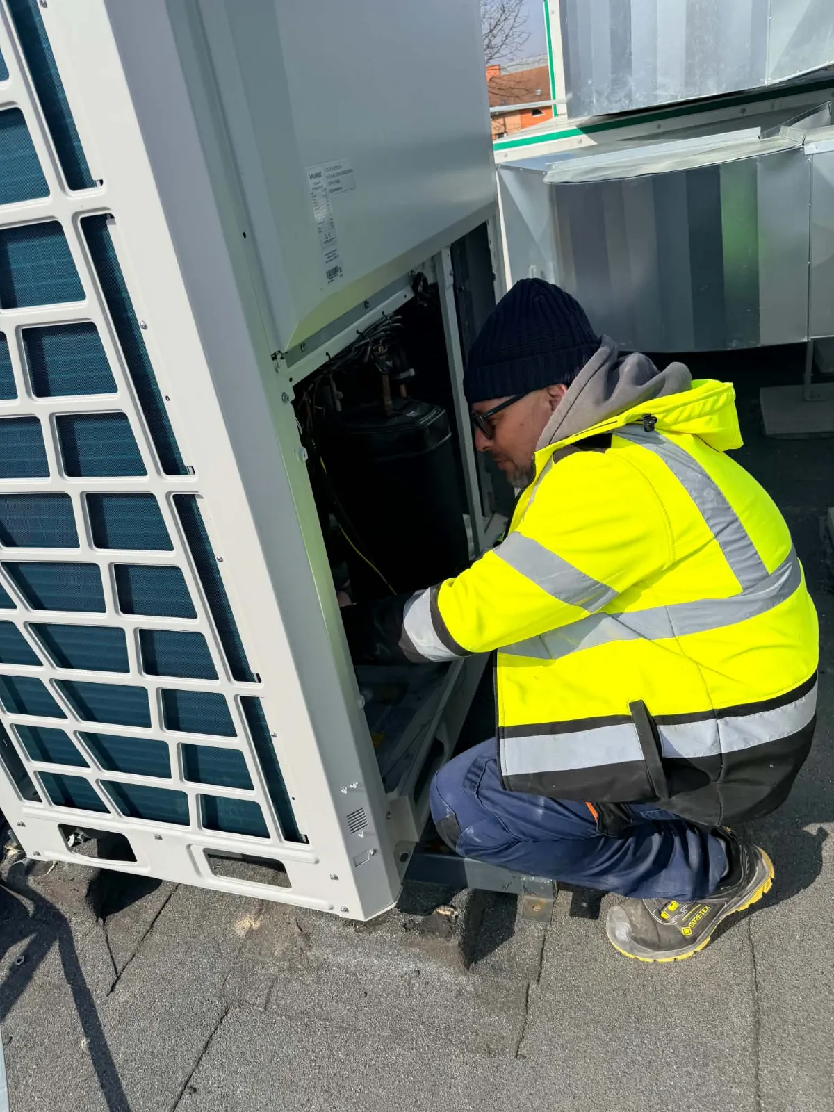 Tehnician îngenuncheat lângă o unitate exterioară de climatizare cu panoul lateral deschis, pe un acoperiș